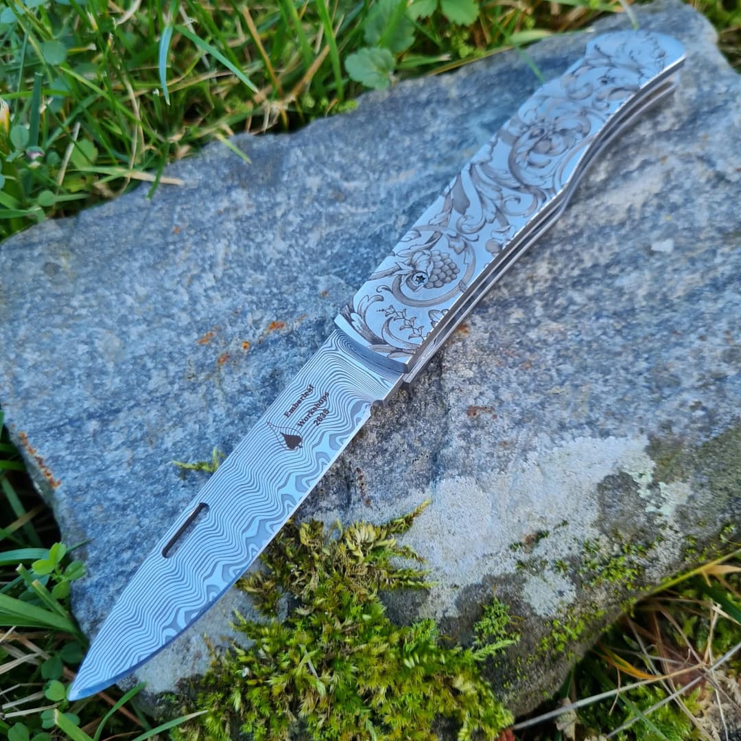 An Emberleaf Aquila lock back folding knife, open, with a damascus blade and an engraved steel handle, on a slab of slate