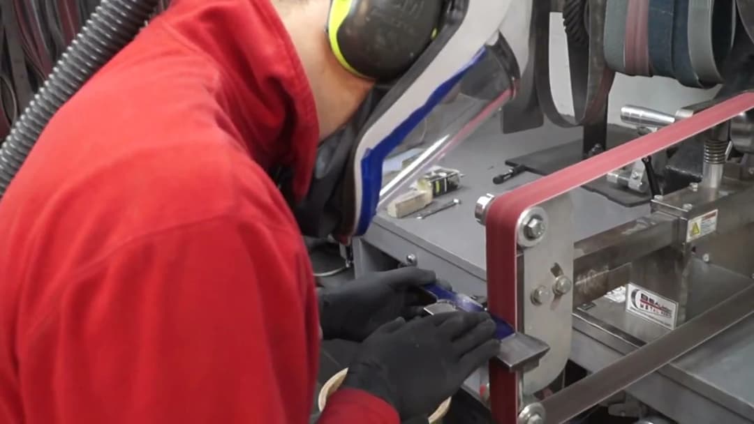 Bladesmith in red jacket and PAPR helmet hand-grinding a taped blade against the belt grinder