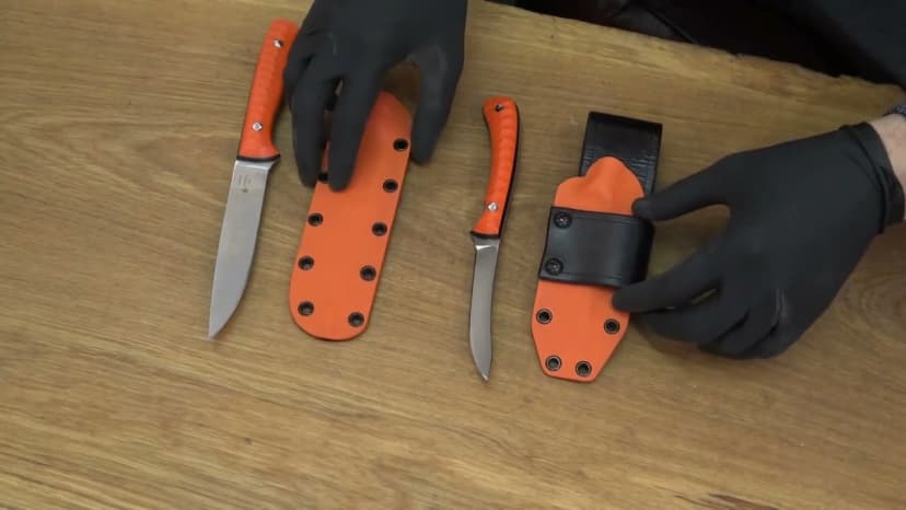 Hybrid Kydex + leather sheath system: orange Kydex shells with removable black leather belt-hangers, two finished knives on oak bench